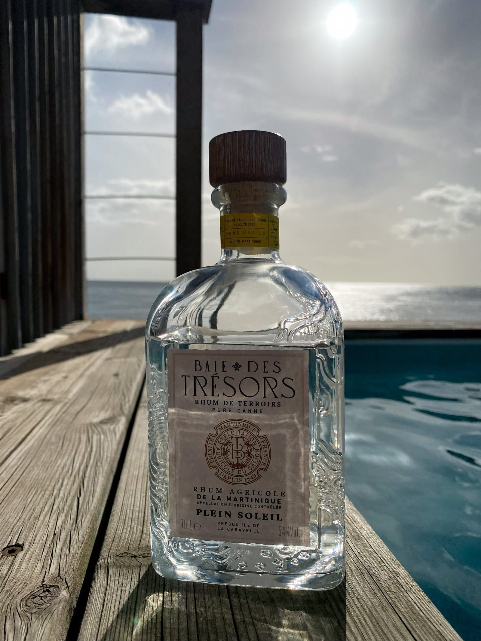 Une bouteille de rhum agricole blanc Baie des Trésors Plein Soleil posée sur une terrasse en bois, face à la mer.