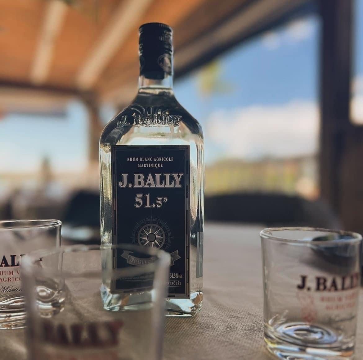 Une bouteille de rhum agricole blanc J. Bally à 51,3°, posée sur un comptoir avec deux verres à l'effigie de la marque.