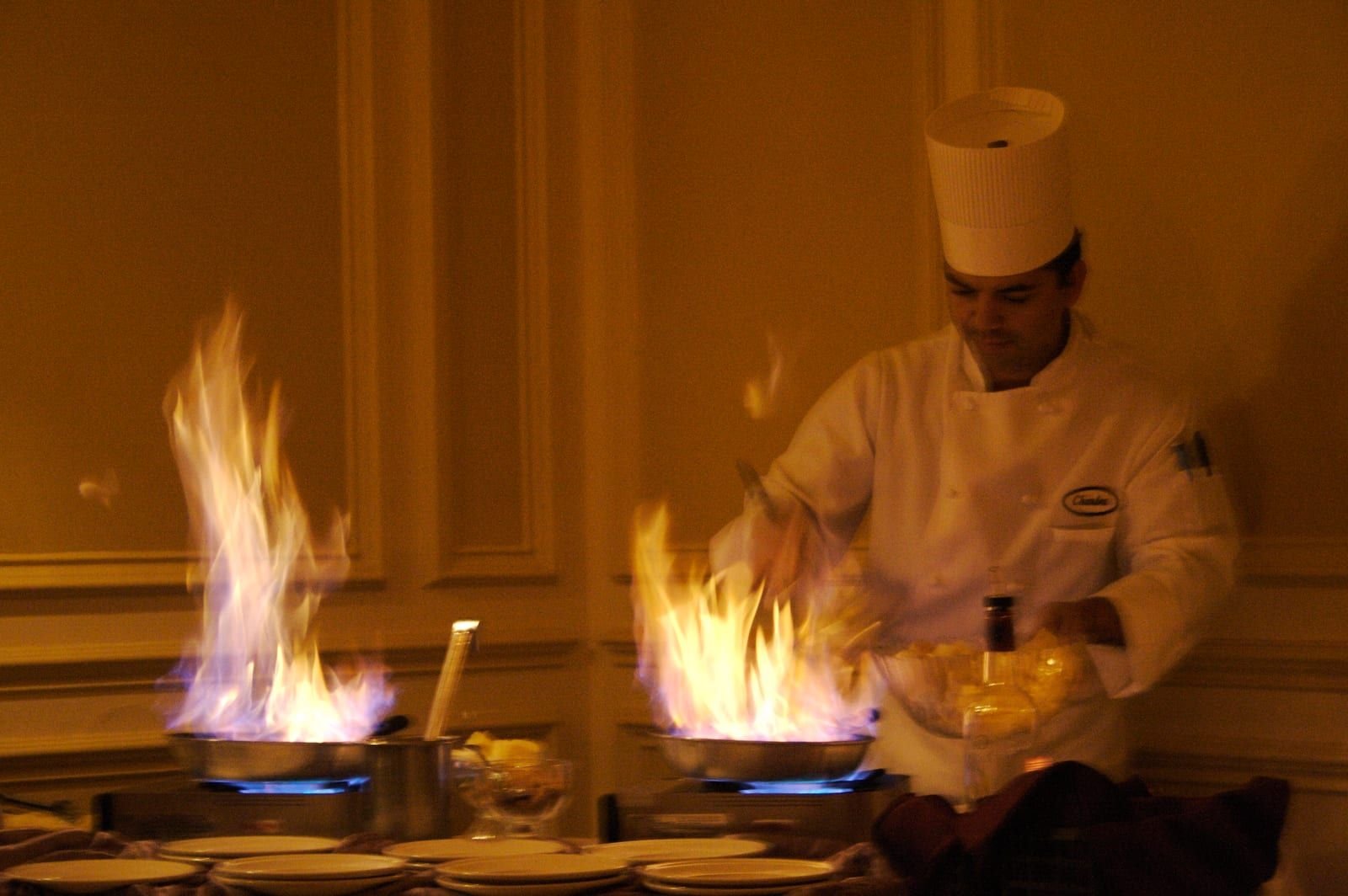 Un cuisinier flambant des crêpes dans deux poêles, hautes flammes orangées devant une pile d'assiettes.