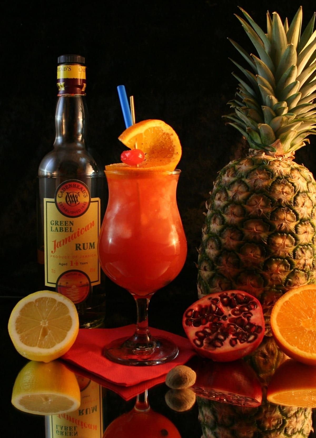 Un punch orangé dans un verre à pied, garni d'une tranche d'orange et d'une cerise, entouré d'un ananas, de fruits coupés et d'une bouteille de rhum.