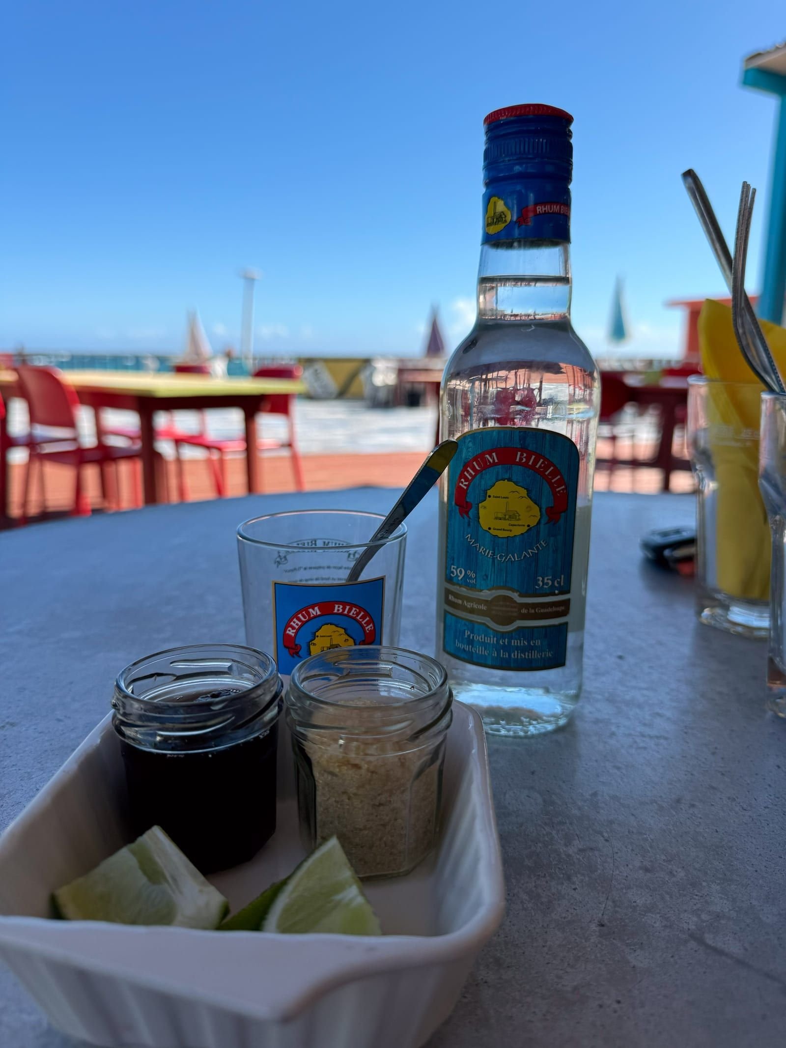 Un ti-punch et une bouteille de rhum blanc sur la table d'un bar de plage, avec des quartiers de citron vert et la mer en arrière-plan.