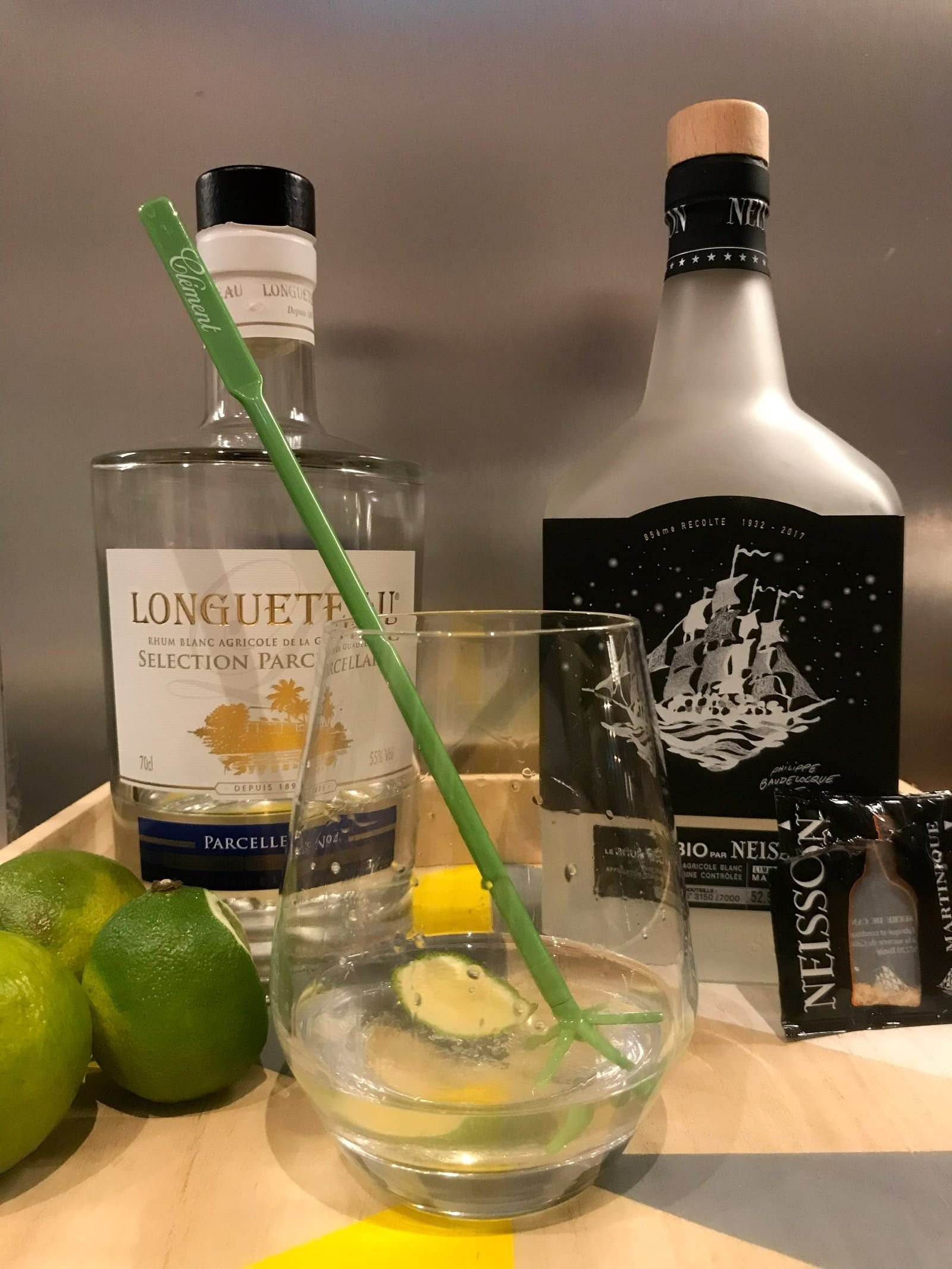 Un ti-punch en préparation : un verre bas contenant une rondelle de citron vert et un bâtonnet, entouré de citrons verts frais, devant une bouteille de Longueteau Sélection Parcellaire et une bouteille Neisson.